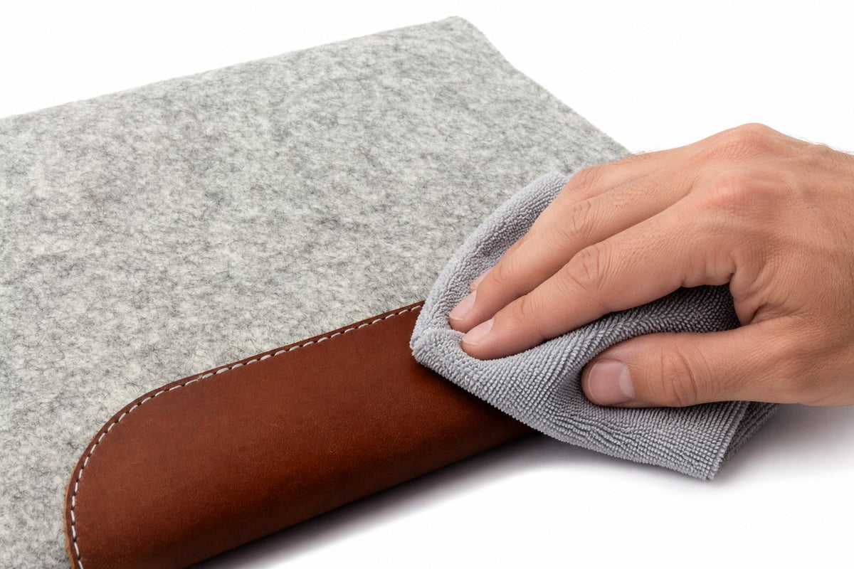 Hand nimmt mit einem weichen Tuch Staub von der Lederlasche einer MacBook Hülle aus Merino-Wollfilz auf