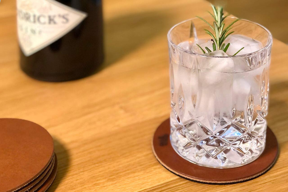 Glas mit Eis und Rosmarin auf einem hellbraunen Glasuntersetzer aus Leder, Holztisch und Ginflasche, Modell DEXTER